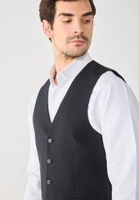 Gilet noir avec cinq boutons, coupe ajustée, tissu lisse, design en V, associé à une chemise blanche à col, poche latérale visible.