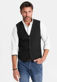Uomo con capelli corti e barba indossa un gilet nero con bottoni sopra una camicia bianca con le maniche arrotolate e jeans blue, con una mano in tasca.