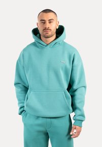 Hoodie turquoise en tissu doux, doté d'une poche avant, d'une capuche à cordon réglable et d'un logo sur la poitrine. Design simple et ample.