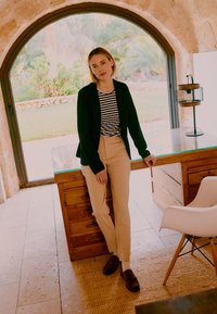 Femme en chemise rayée, veste noire et pantalon beige, appuyée sur un bureau en bois dans une pièce lumineuse avec une fenêtre en arc et une chaise blanche à proximité.