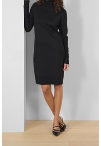 Svart stickad turtleneckklänning med långa ärmar, ribbade detaljer vid ärm- och nederkanter, tillsammans med spetsiga svarta klackar och ett slätt trägolv.