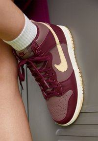 Vysoké tenisky z bordové kůže s krémovými doplňky. Mají perforovanou špičku a hnědou gumovou podrážku. Šněrování a logo jsou výrazně viditelné.