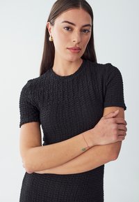 Schwarzes, strukturiertes Kleid mit kurzen Ärmeln und rundem Ausschnitt. Das Model posiert mit verschlossenen Armen und zeigt subtile rote und grüne Handgelenkstattoos.