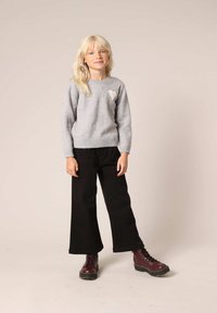 Grijze gebreide trui met een hartdetail aan de linkerkant, gecombineerd met zwarte wijde broekspijpen en bordeauxrode veterschoenen.