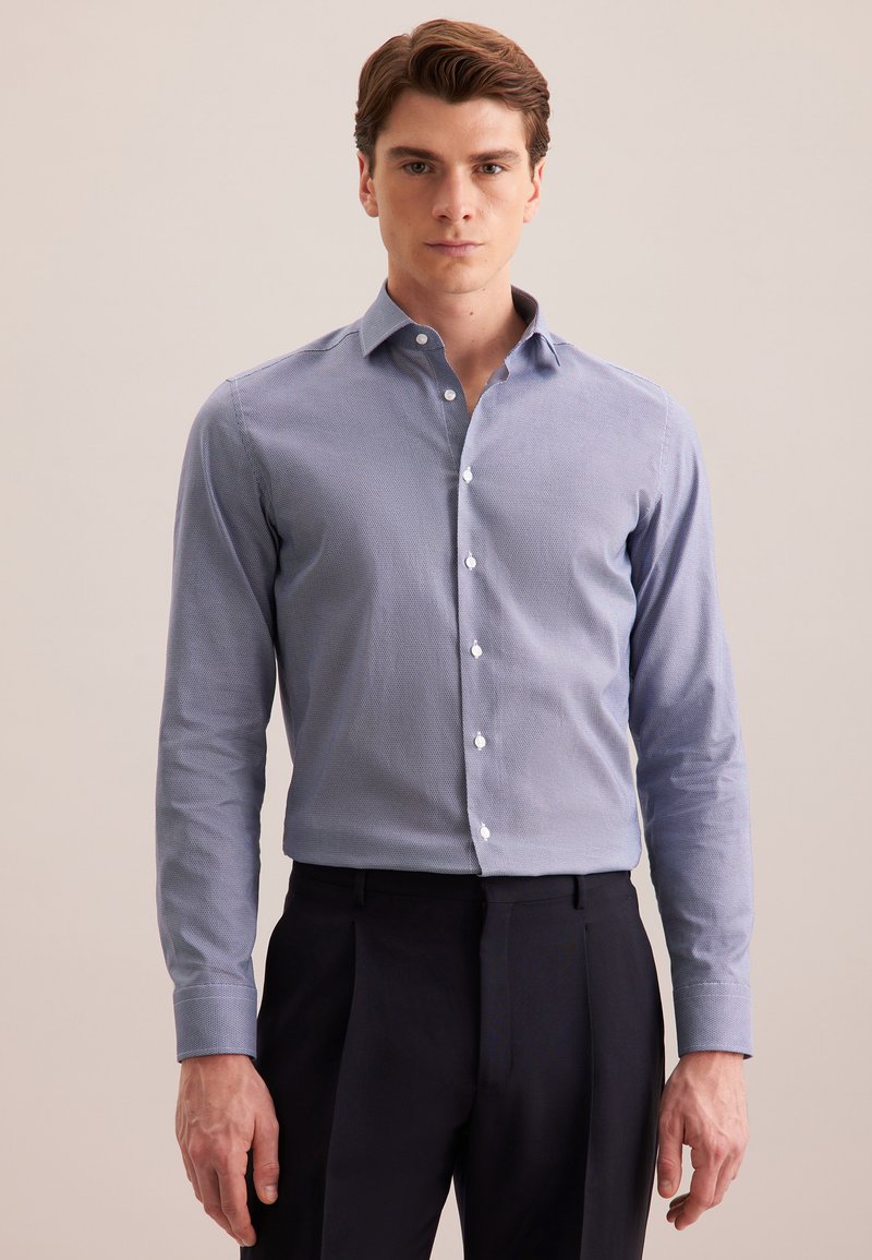 Man in een lichtblauw, gemusterd, langemouw overhemd, ingestopt in een zwarte pantalon, staande tegen een effen beige achtergrond.