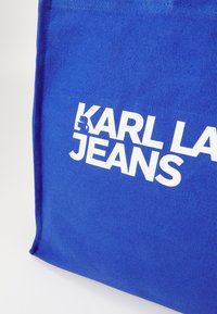 Mėlyna drobės pirkinių rankinė su baltais užrašais "KARL LAGERFELD JEANS" vienoje pusėje. Audinys turi tekstūruotą paviršių ir tvirtą siuvimą.