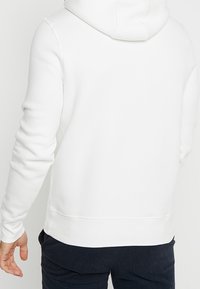 Bílá bavlněná mikina s kapucí na stahovací šňůrku, žebrovanými manžetami a lemem; má hladkou texturu a jednoduchý, nenápadný design.