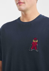 Camiseta de algodón azul marino con un pequeño diseño de esquiador bordado en rojo en el lado izquierdo del pecho. Cuello crew clásico y mangas cortas.