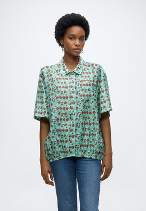 Femeie cu păr scurt natural, purtând o cămașă turcoaz largă cu nasturi și model de căpșuni roșii și blugi albaștri, stând nemișcată.