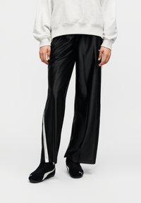 Άτομο που φοράει μαύρο παντελόνι με φαρδύ πόδι και λευκή ρίγα στο πλάι, λευκό φούτερ και μαύρα αθλητικά Puma, στέκεται πάνω σε λευκό φόντο.