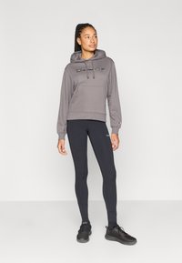 Γκρι φούτερ με κουκούλα, κορδόνια, εμπρόσθια τσέπη και λογότυπο· συνδυασμένο με μαύρα κολάν και μαύρα αθλητικά παπούτσια. Ομοιόμορφη υφή σε όλο το ύφασμα.