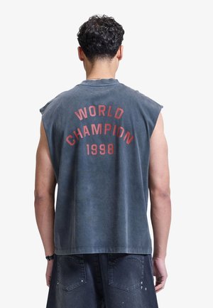 Uomo con capelli neri ricci che indossa una maglietta senza maniche grigio scuro con scritta rossa "Campione del Mondo 1998" stampata sulla schiena e pantaloncini in denim neri.