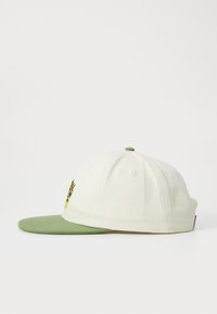 Hvit bomullslue med grønn visor, som har et gult grafisk design på forsiden. Inkluderer øyehull og en toppknappdetalj.