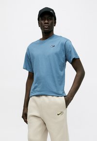 Άνδρας με μαύρο καπέλο Nike, μπλε μπλουζάκι Nike και μπεζ φόρμα Nike, στέκεται με το ένα χέρι στην τσέπη μπροστά σε λευκό φόντο.