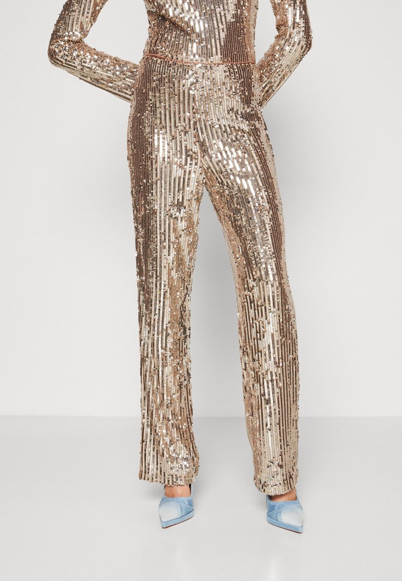 Mujer con pantalones anchos de lentejuelas doradas y top a juego, combinados con zapatos de tacón puntiagudo azul claro, de pie frente a un fondo liso.