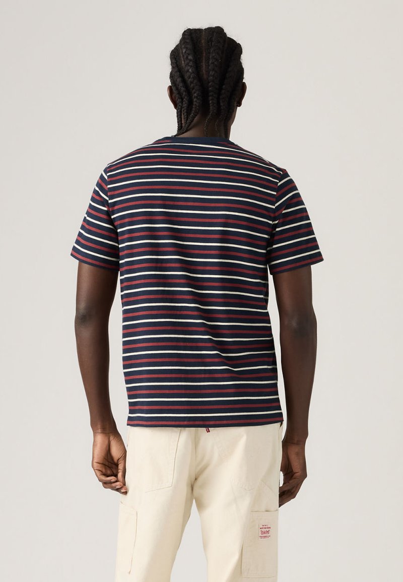 Person mit geflochtenem Haar, der ein kurzärmeliges Hemd mit Marineblau-, Rot- und Weißstreifen sowie helle beige Hose trägt und mit dem Rücken zum Betrachter vor einem einfarbigen Hintergrund steht.