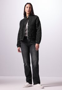 Chaqueta negra acolchada con cuello alzado, botones a presión y dos bolsillos frontales, combinada con jeans de mezclilla negros y zapatillas blancas.