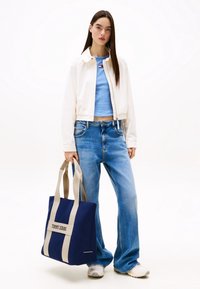 Blå tygväska med ljusbeige handtag, med en "TOMMY JEANS"-etikett. Bärare med en krämfärgad jacka och en ljusblå topp, ihop med blå jeans.