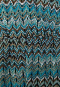 Tejido de punto con un patrón en zigzag en tonos de teal, marrón y blanco, que presenta calados y diversas texturas a lo largo.