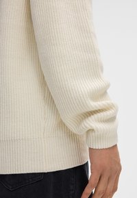 Nahaufnahme einer Person, die einen cremafarbenen gerippten Pullover und dunkle Hosen trägt, wobei die Strickstruktur des Pullovers und das Detail der Bündchen sichtbar sind.
