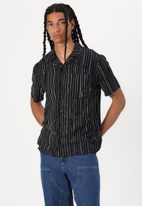 Short-sleeve black shirt featuring vertical white stripes, a collar, a left chest pocket, and a relaxed fit. Made from lightweight fabric.