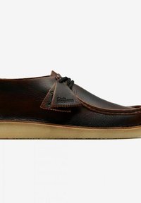 Scarpa in pelle marrone con forma arrotondata, caratterizzata da un accento cucito, una patch in pelle e suola in gomma. I lacci sono integrati nella parte superiore.