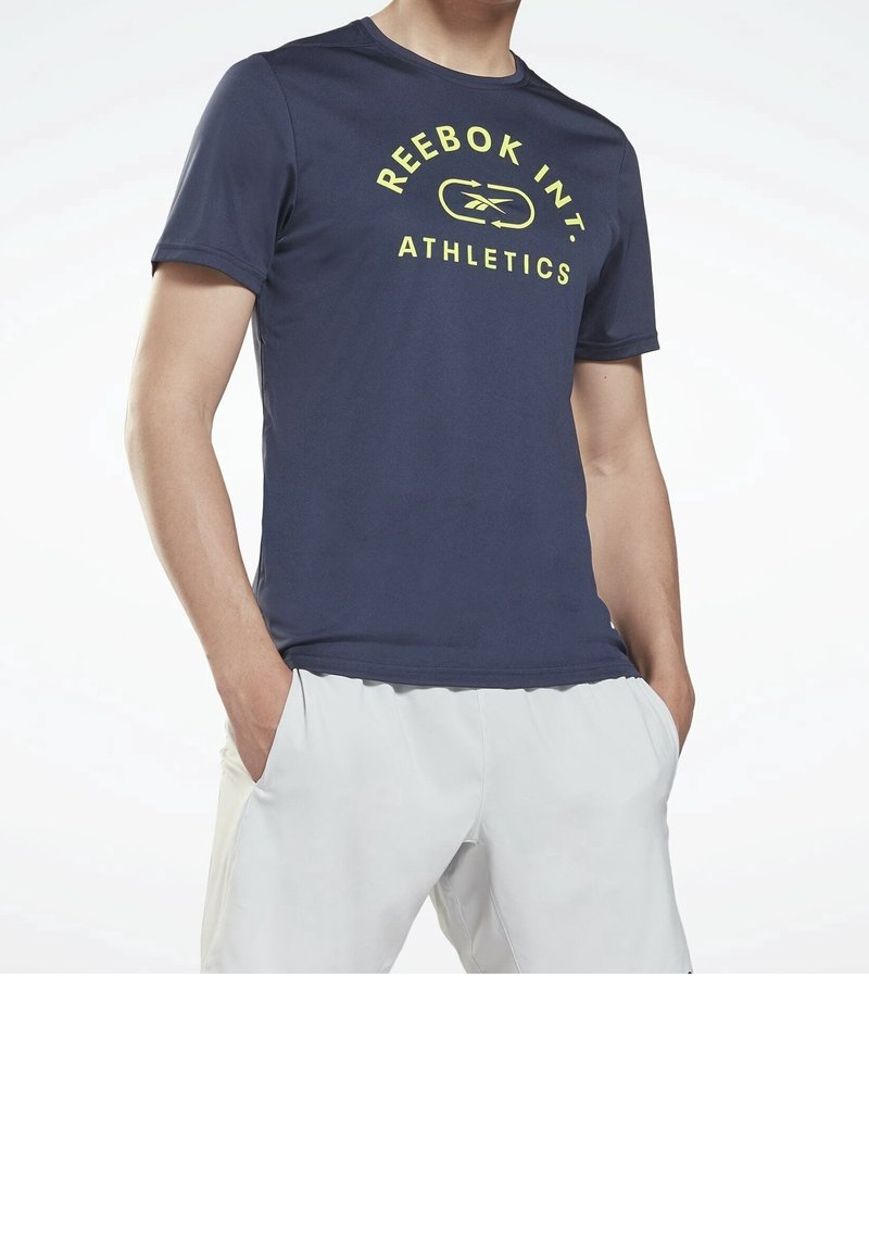 Homme portant un t-shirt bleu marine Reebok avec un logo jaune et un short blanc, les mains dans les poches, sur un fond clair uni.