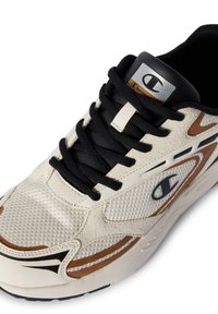 Beige Sneaker mit schwarzen Schnürsenkeln, strukturierten Mesh-Panels, braunen Akzenten und einem silbernen Logo auf der Zunge. Rundliche Zehenpartie und gepolsterte Sohle.