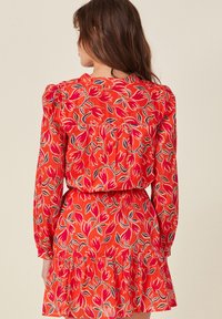 Punainen kukallinen mekko, jossa on pitkät hihat, kerätyt olkapäät, kerroksellinen röyhelöhelma sekä pinkkien ja mustien tulppaanien kuvio kirkasta taustaa vasten.