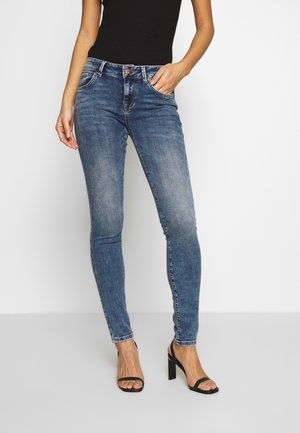 Woman wearing fitted blue denim jeans and black heeled sandals, hand resting in front pocket against white background.