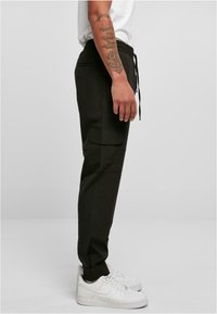 Zwarte cargo broek met een trekkoord in de taille en twee zijzakken, gecombineerd met witte sneakers. Het materiaal lijkt zacht en heeft een relaxte pasvorm.