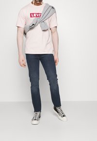 Camiseta de algodón rosa claro con el logo rojo de "LEVI'S", combinada con vaqueros ajustados azul oscuro y zapatillas altas en blanco y negro. Sudadera gris caída sobre los hombros.