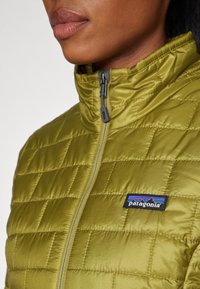 Oliven grønn, quiltet pufferjakke med høy krage og glidelås. Har en svart Patagonia-logo med blå og røde detaljer.