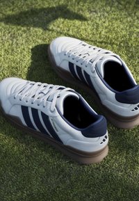 Ζευγάρι λευκά και ναυτικά μπλε αθλητικά παπούτσια με λαστιχένιες σόλες σε επιφάνεια πράσινου χόρτου, από ψηλά σε γωνία.