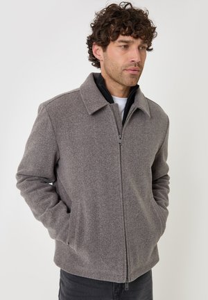 Hombre con pelo rizado que lleva una chaqueta de lana gris con cremallera y las manos en los bolsillos, mirando ligeramente hacia un lado.