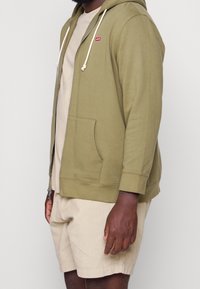 Hombre vistiendo una sudadera olive verde con cremallera y cordones blancos, una camiseta beige y pantalones cortos beige, de pie contra un fondo blanco.