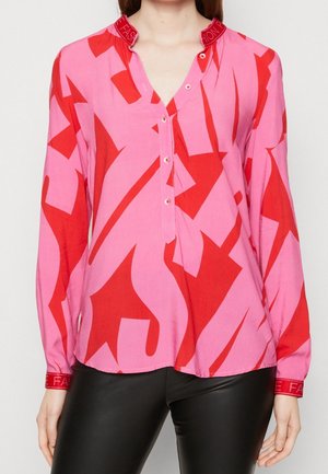Blusa rosa de manga larga con patrones abstractos rojos audaces, cierre de botones en el frente y cuello y puños rojos con texto blanco, combinada con pantalones negros.