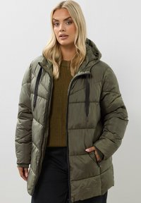 Doudoune vert olive avec capuche, présentant un design matelassé, une fermeture zippée et des accents à cordon. Tissu texturé et poches latérales.