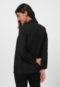 Blazer noir avec une coupe ajustée, des manches longues et un col à revers. Comprend une poche avant unique et un tissu lisse et texturé.