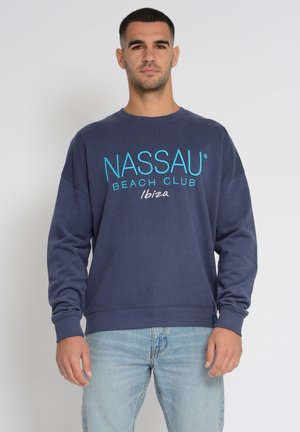 Mørkeblå sweatshirt med "NASSAU BEACH CLUB Ibiza" grafik i turkis og hvid. Har ribbede manchetter og afslappet pasform. Bæres med lyseblå jeans.