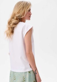 Witte top met korte mouwen, een relaxte pasvorm en een ronde halslijn. Opmerkelijke details zijn een label aan de zijkant en een gladde textuur.