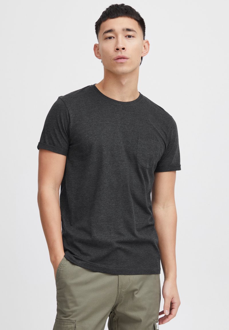 T-shirt à manches courtes gris foncé avec un col rond et une poche sur la poitrine. Texture douce. Associé à un pantalon cargo vert olive.