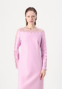 Vestido rosa de manga larga con escote y mangas con borde ondulado, paneles transparentes, textura de tela suave y silueta ajustada.