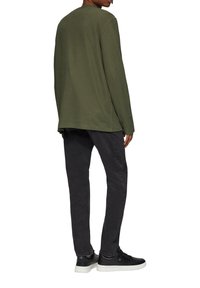 Olivgrünes Langarmshirt mit abgerundetem Saum, kombiniert mit schwarzen Slim-Fit-Hosen und schwarzen Turnschuhen mit weißen Sohlen.