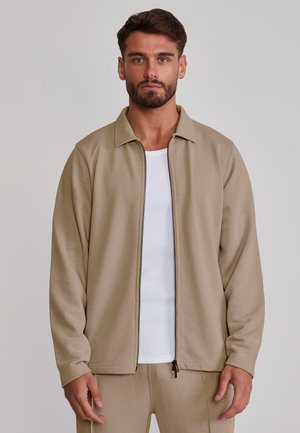 Mann mit Bart trägt eine beige Reißverschlussjacke über einem weißen Hemd, steht vor einem schlichten hellen Hintergrund, schaut nach vorn.