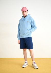 Light blue hoodie with drawstring and front pocket, navy shorts, pink cap, and multi-colored chunky sneakers on a yellow floor.