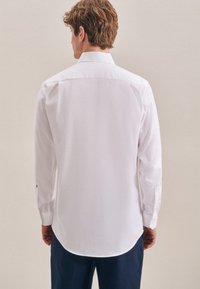 Camicia bianca a maniche lunghe con colletto classico, orlo curvo e polsini abbottonati. Tessuto liscio con una leggera lucentezza, vestibilità sartoriale.