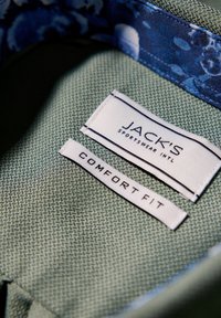 Tessuto verde texturizzato con due etichette: "JACK'S SPORTSWEAR INTL" in nero e "COMFORT FIT" in bianco, situate su uno sfondo floreale blu.