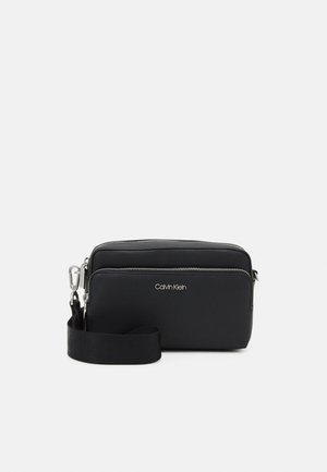 Schwarze rechteckige Calvin Klein Crossbody-Tasche mit zwei Reißverschlussfächern und einem verstellbaren schwarzen Riemen mit Metallverschluss.