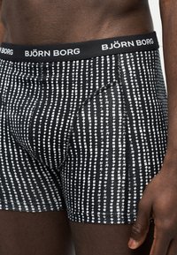 Μπάρξερ εσώρουχα με ασπρόμαυρο σχέδιο και το λογότυπο Björn Borg στη ζώνη μέσης, φορεμένα από ένα άντρα με σκούρο δέρμα, το χέρι του χαλαρό στο πλάι.